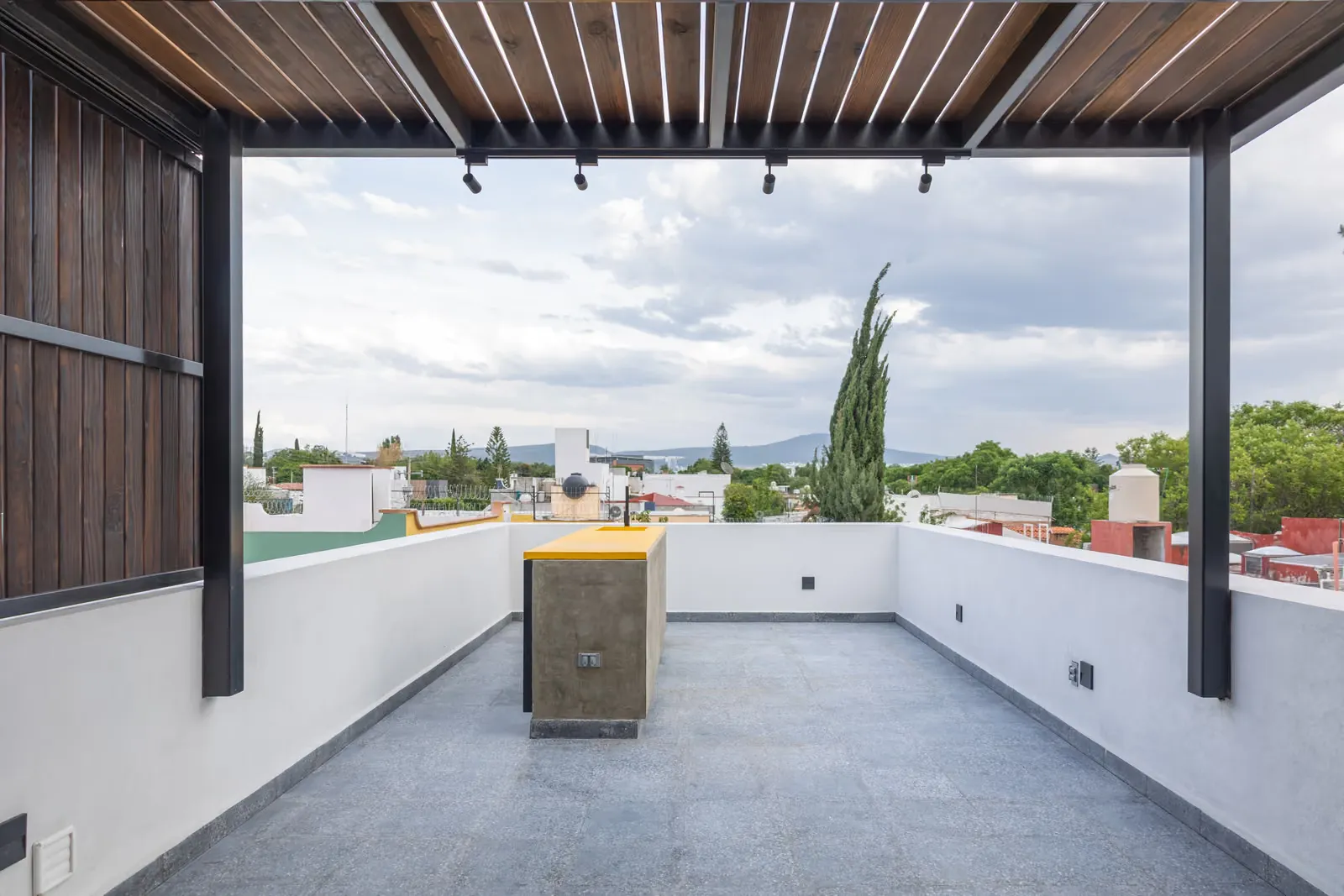 Azotea de Casa Seto con pretiles blancos, piso gris y una barra de concreto bajo una pérgola de madera, con árboles y casas vecinas al fondo